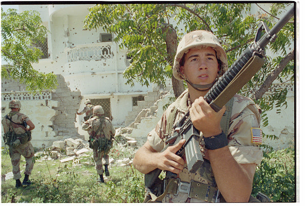 Jarrett holding a rifle in a combat zone "Somalia" with other soldiers in the background.